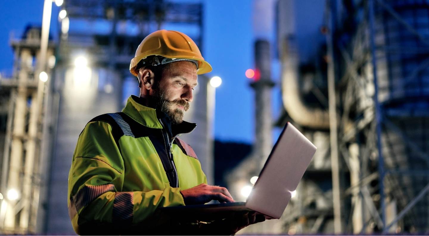 Photo of an engineer looking at a laptop with a chemical plant in the background